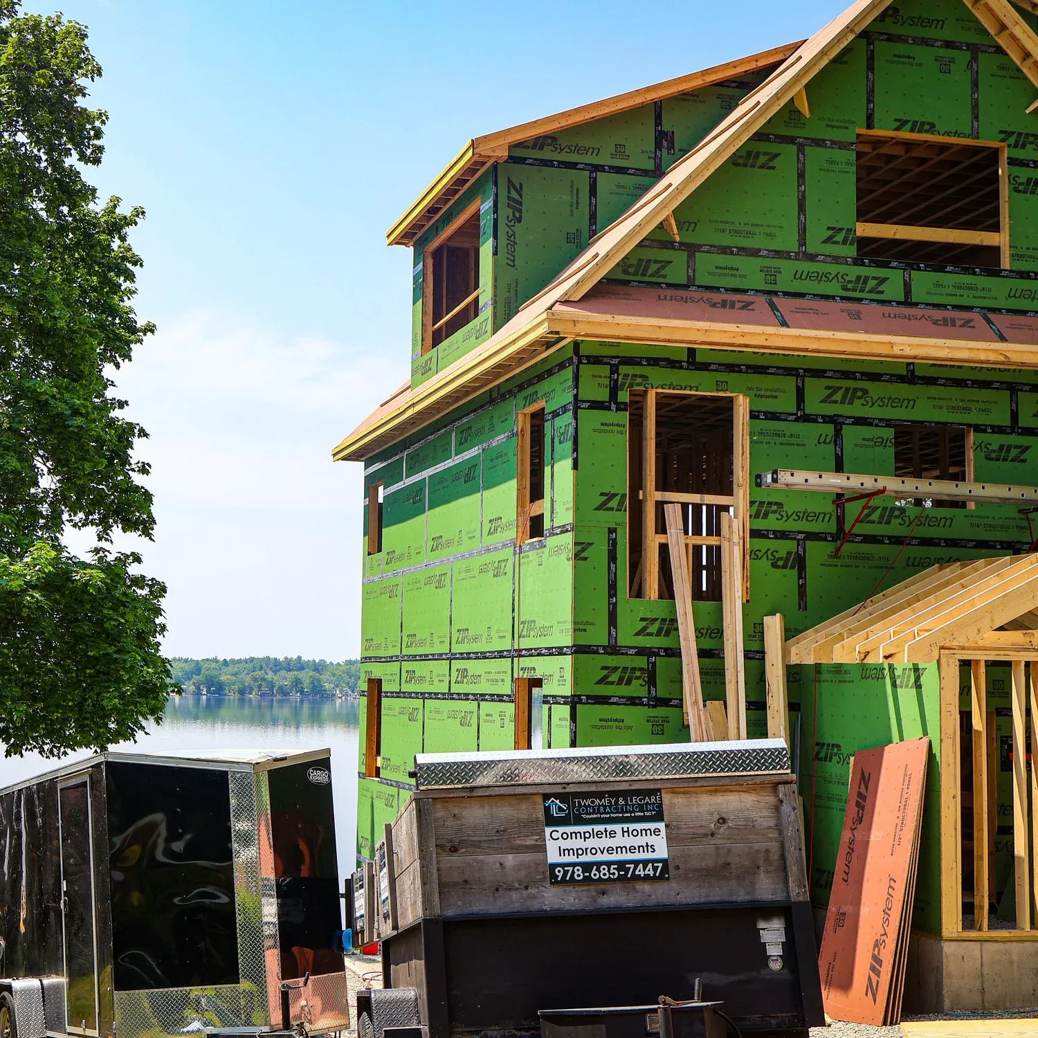 A house under construction with green insulation sheathing at a lakeside, with trees and water in the background, and construction equipment and materials in the foreground.
