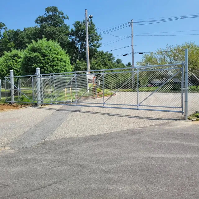 Chain-link fence gate, closed, on gravel drive. Trees and power lines in the background on a sunny day.