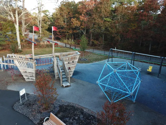 Empty children's playground with wooden ship-shaped climbing structure, blue geometric climbing frame, yellow swing, and bench, surrounded by trees with autumn foliage.