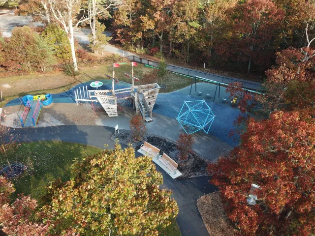 aerial view of a playground surrounded by autumn trees with red, orange, and green leaves, featuring a wooden climbing structure, swings, a merry-go-round, and benches.