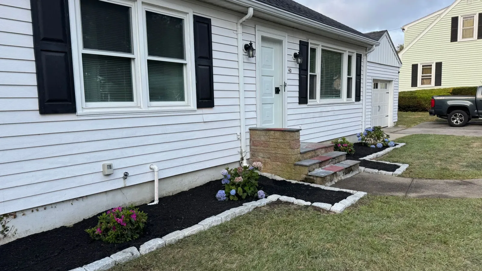 White house with black shutters, door, and a landscaped front yard with flowers and dark mulch.