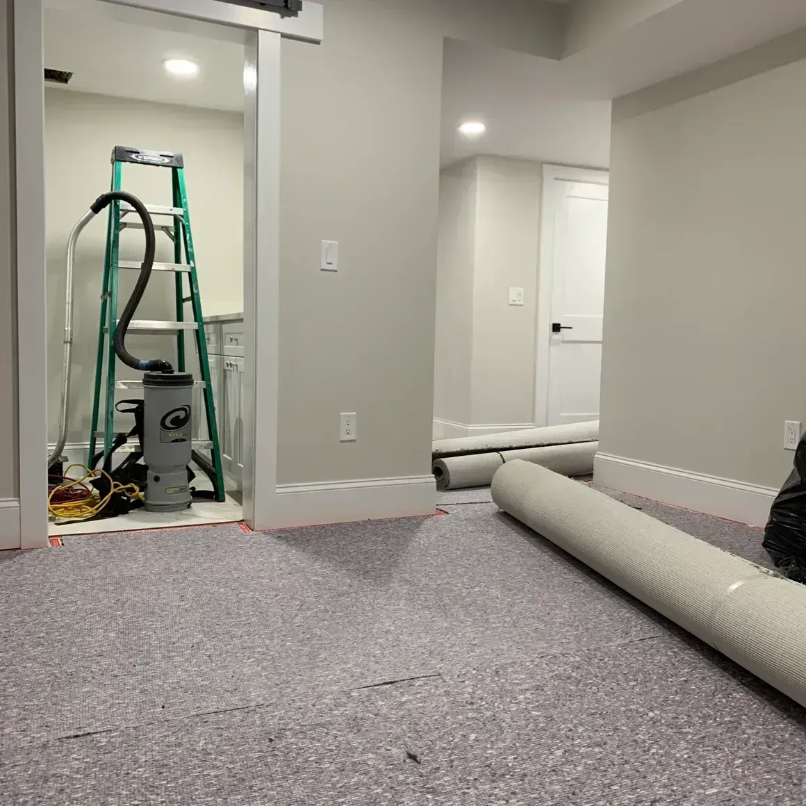 Basement with new flooring being installed; rolled carpet, ladder, vacuum.