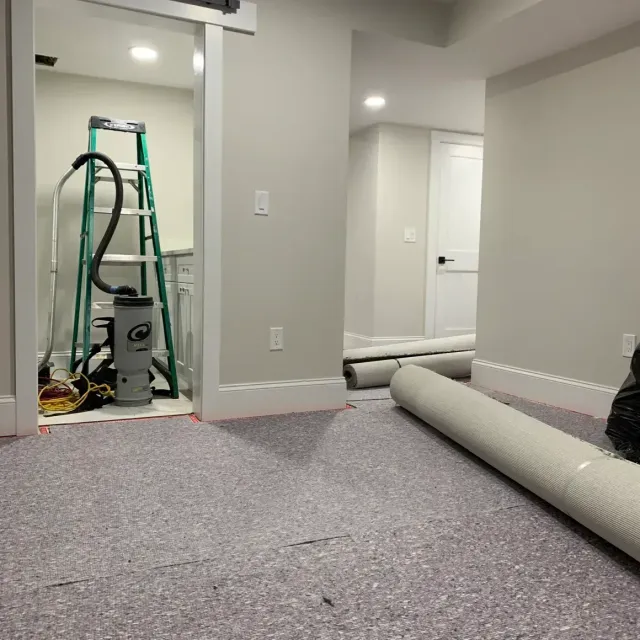 Basement with new flooring being installed; rolled carpet, ladder, vacuum.