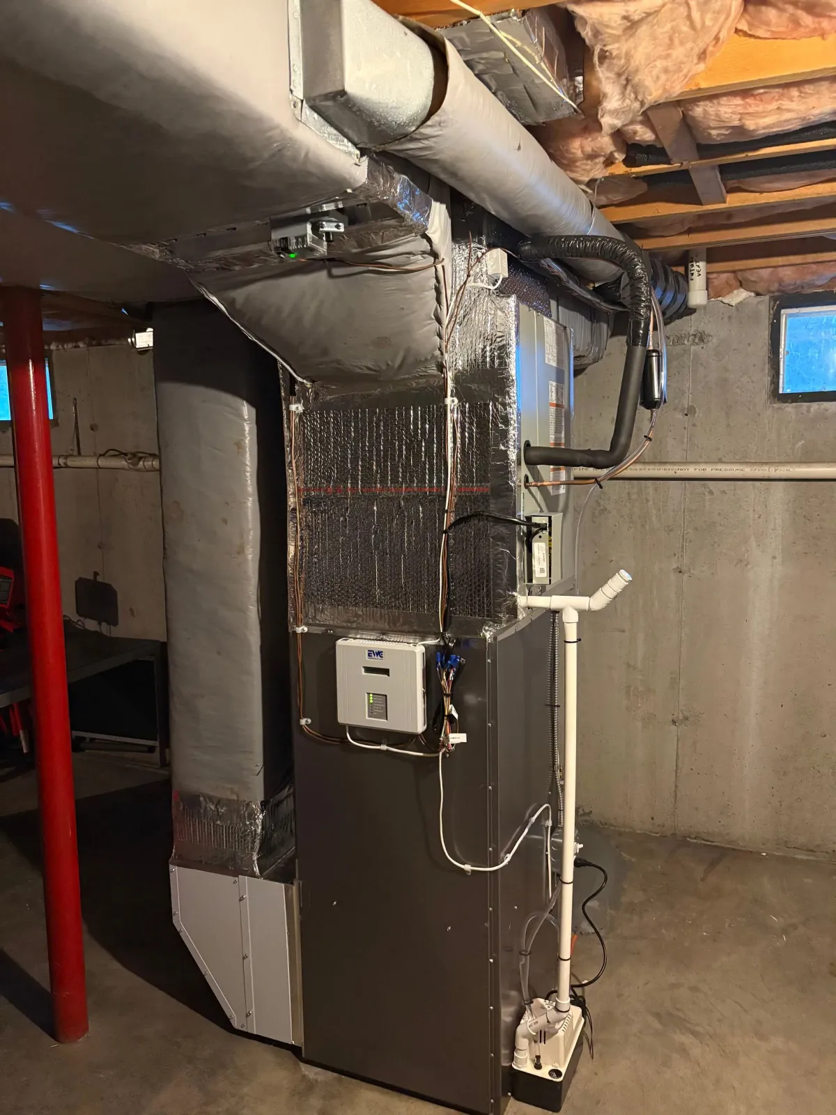 Furnace and ductwork in a basement. Grey metal unit with connected silver ducting. Concrete walls, support pole.