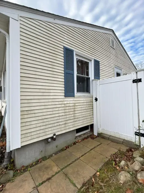 Light beige house exterior with blue shutters, a small basement window, white fence gate, and stone path beside grassy area and rocks.