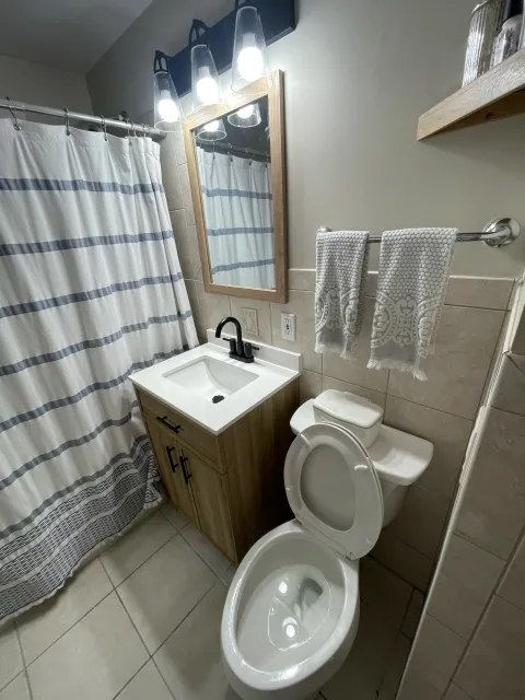 Bathroom with a shower curtain, mirror with wooden frame, light fixture above the mirror, towels on a silver towel rack, a small wooden vanity with a black faucet, and a white toilet with the lid open. There are beige tile walls and floor.