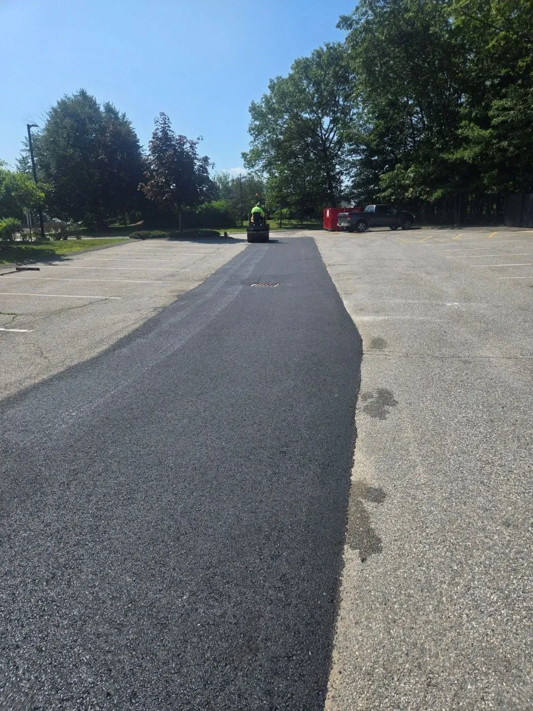 Freshly paved dark asphalt path through a sunny park with trees and a person in red in the distance
