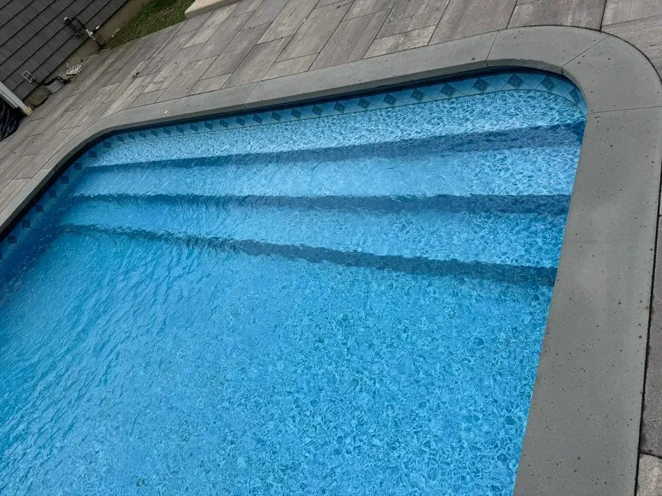 A set of built-in swimming pool stairs with a light blue patterned liner, bordered by a dark concrete coping and deck.