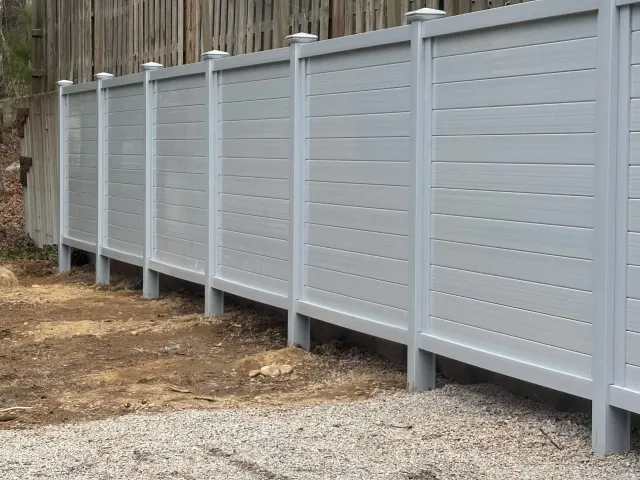 Gray horizontal slat fence in front of a wooden fence, on a gravel surface.