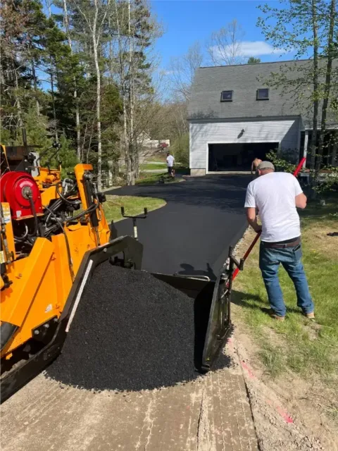 Asphalt paving a driveway. A worker shovels fresh blacktop. An orange paver machine is also in use.