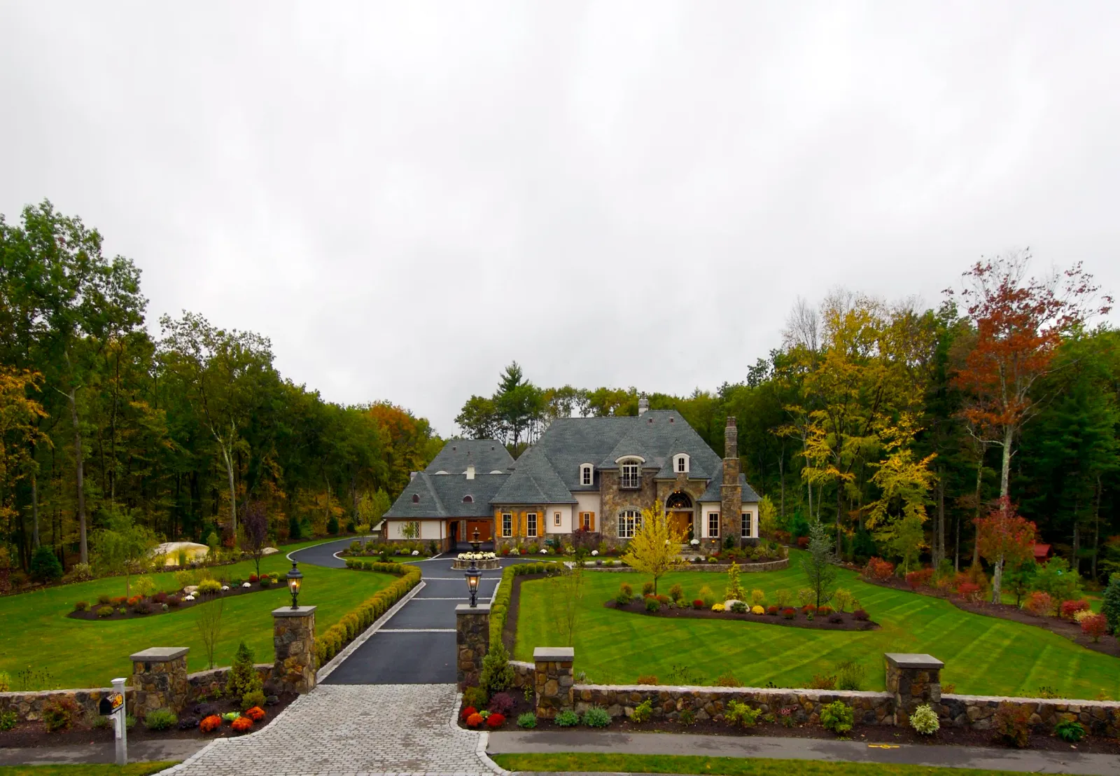 Large house with stone facade, located in a well-maintained front yard with green grass, flower beds, paved driveway, and surrounded by trees with fall foliage.