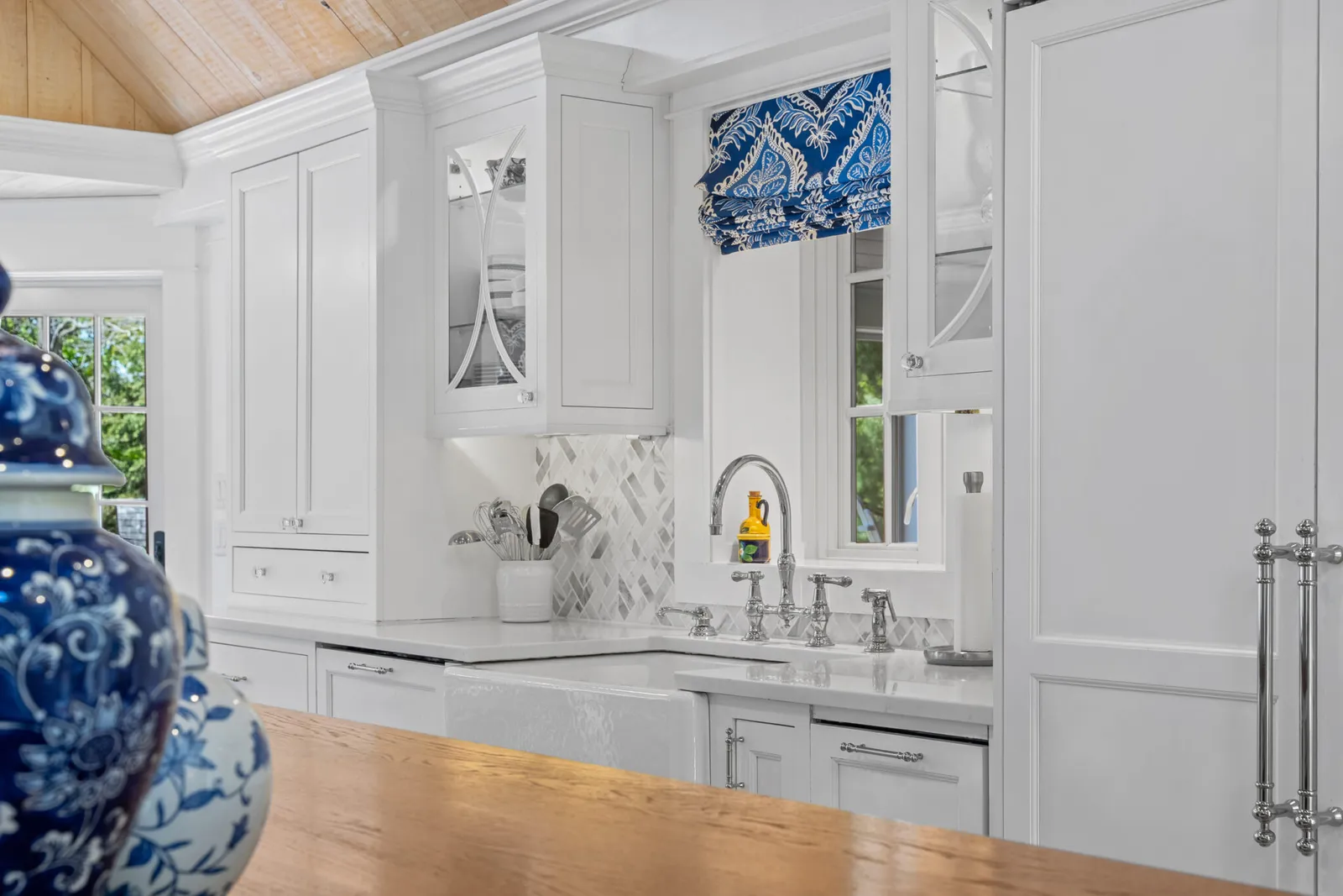 Close-up of a custom kitchen sink area with white inset cabinetry, apron farmhouse sink, polished chrome bridge faucet, white quartz countertops, diamond herringbone marble tile backsplash, and glass-front upper cabinets