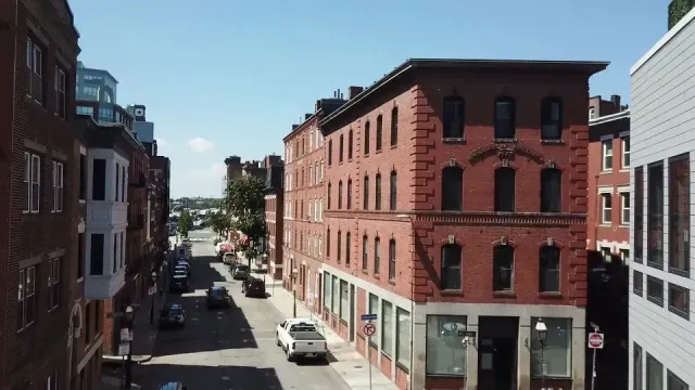 Brick buildings line a sunny city street; several cars drive down the road.