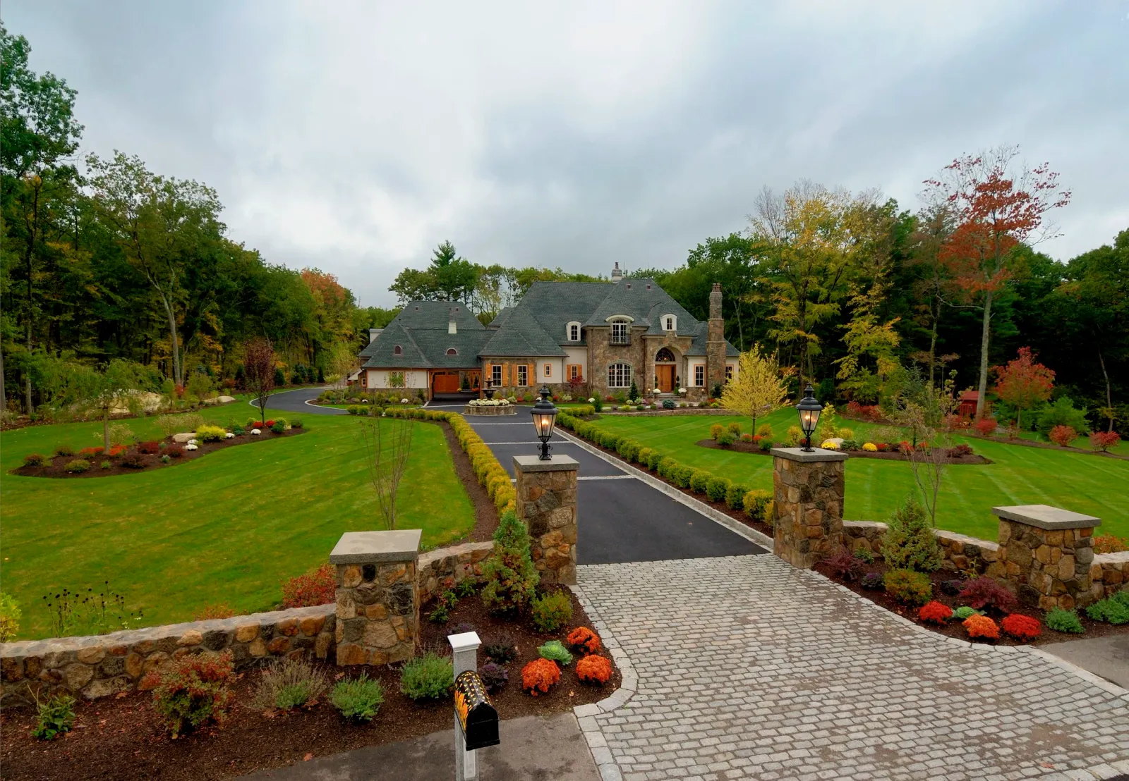 A large house with a dark gray roof and a stone exterior, surrounded by a landscaped front yard with a cobblestone driveway, brick pillars with lanterns, flower beds with colorful plants, and a well-maintained green lawn, trees, and bushes in the bac