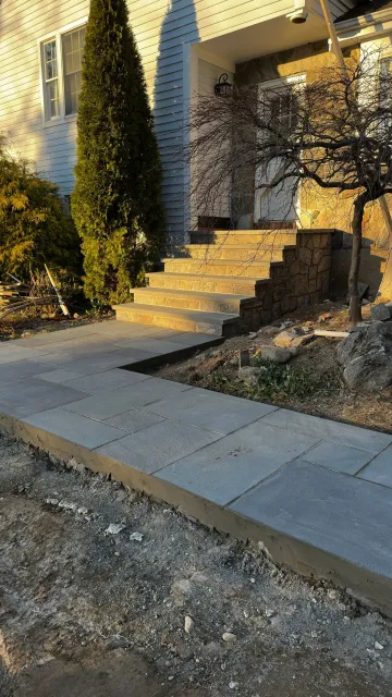 Front steps with stone railing beside a house, lit by warm sunlight, with a sidewalk and gravel foreground.