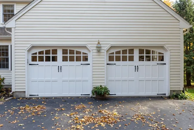 White double garage doors on a suburban house with white siding and a leaf-covered driveway.