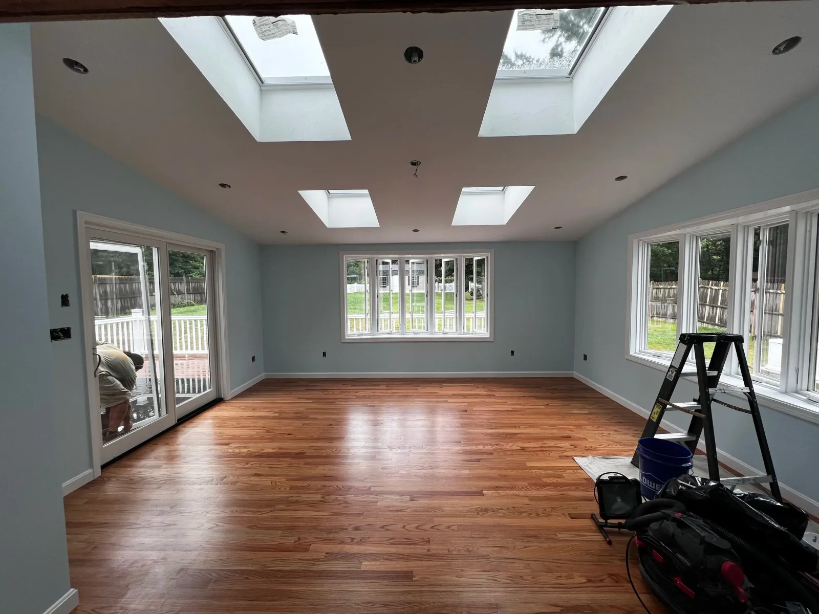 Empty living room with new hardwood floors, light blue walls, large windows, and skylights, with construction tools and ladder near the right wall.