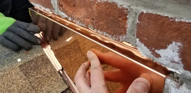 Hands installing copper flashing on brick, next to shingles.