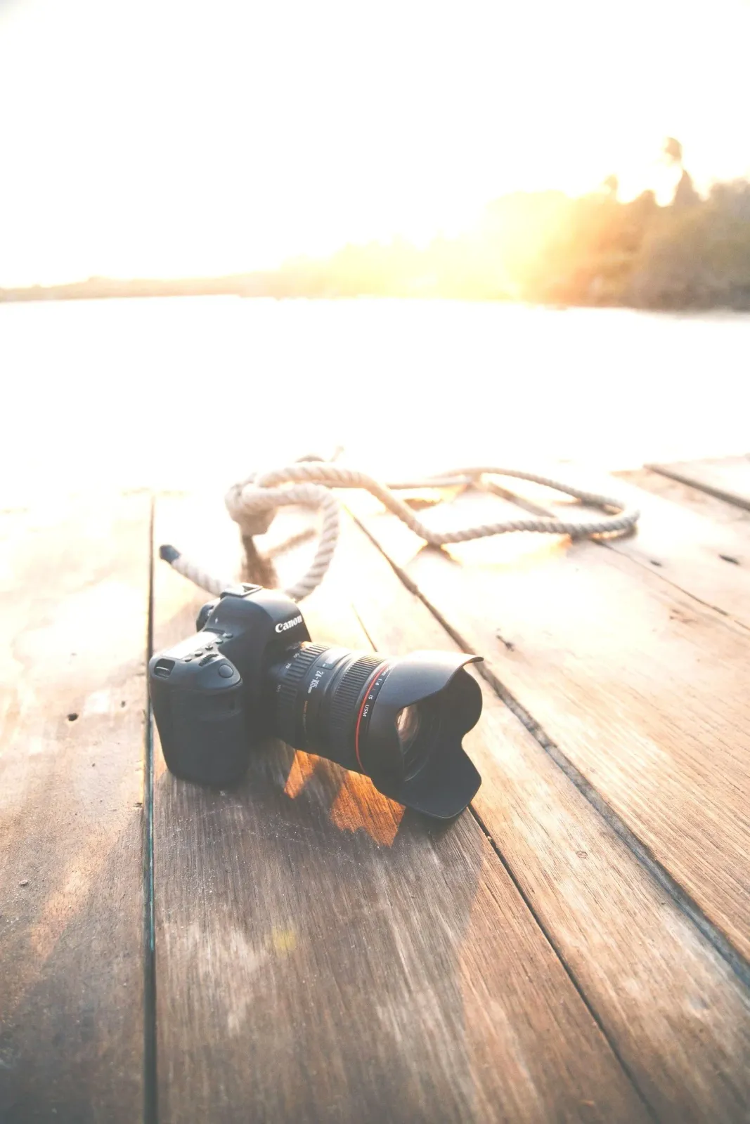 A Canon DSLR camera with a zoom lens and lens hood rests on a wooden dock near a body of water, illuminated by sunlight from the horizon.