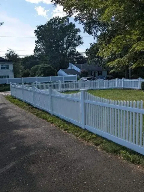 White three-rail fence alongside a paved driveway and green lawn, with trees in the background.
