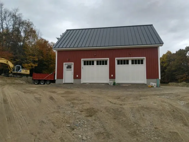Red garage with two white doors, one side door, dark roof, dirt lot, excavator.