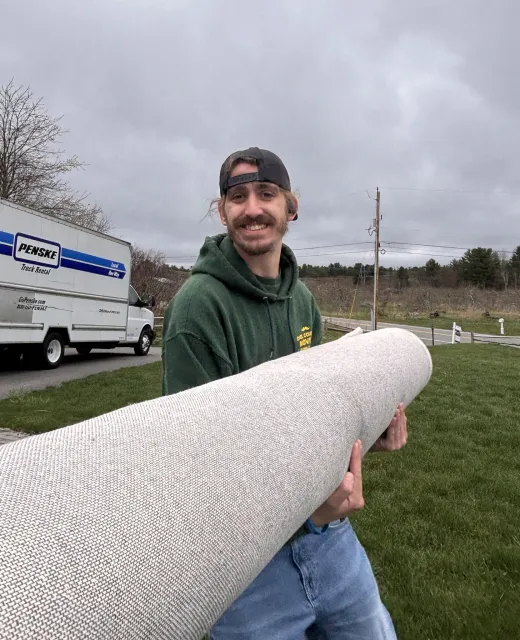 Man holding a rolled white rug outdoors near a moving truck under a cloudy sky.