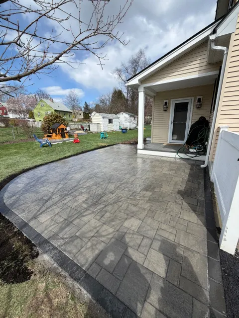 Stone patio beside a white house with a covered porch, overlooking a backyard and blue sky.