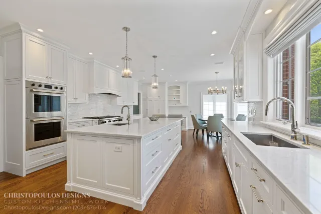 Bright, spacious kitchen with white cabinets and marble countertops. Recessed lighting and pendant lights hang over the island. Large windows with white blinds let in natural light, overlooking green trees.