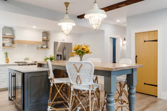 Modern kitchen with a large wooden island, wicker chairs, pendant lights, stainless steel refrigerator, and sunflowers centerpiece. Features a rustic yellow door and open shelving with dishes and pineapples.