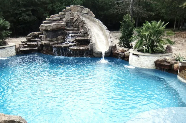 A backyard swimming pool with a small waterfall feature surrounded by rocks and lush green plants.