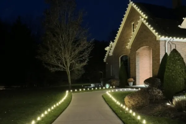 Warm landscape lighting highlights a brick home at night. Path lights illuminate the driveway, leading to the house with roofline lighting, creating a welcoming ambiance.
