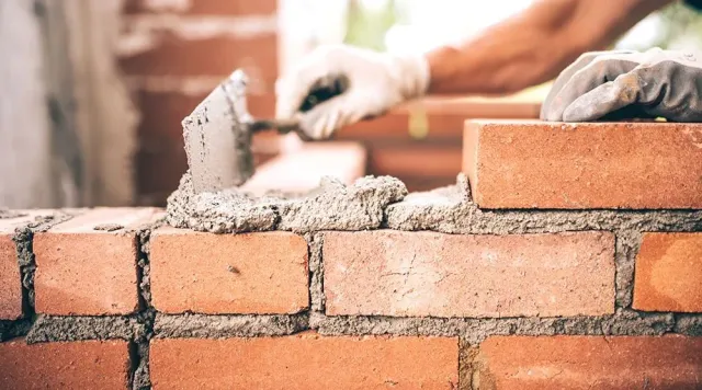 Close-up of gloved hands using a trowel to apply mortar on red bricks, constructing a wall.