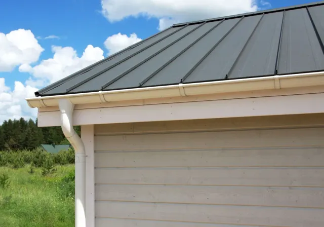 Gray metal roof and white gutter on a beige building under a blue sky with clouds