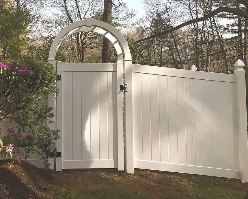 White arched wooden gate and fence beside blooming pink bushes in a wooded yard