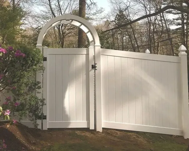 White arched wooden gate and fence beside blooming pink bushes in a wooded yard
