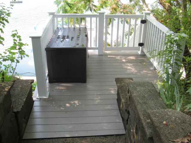 Small wooden deck with white railing and a black storage box, overlooking a body of water surrounded by greenery.