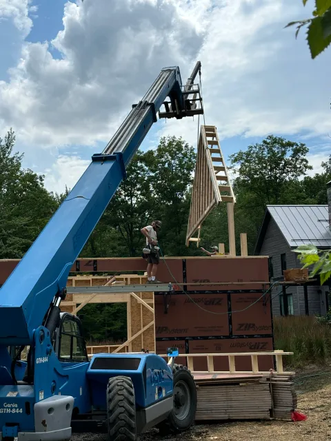 Construction workers are lifting a wooden wall into place on a building under construction, with a large blue crane and trees in the background.
