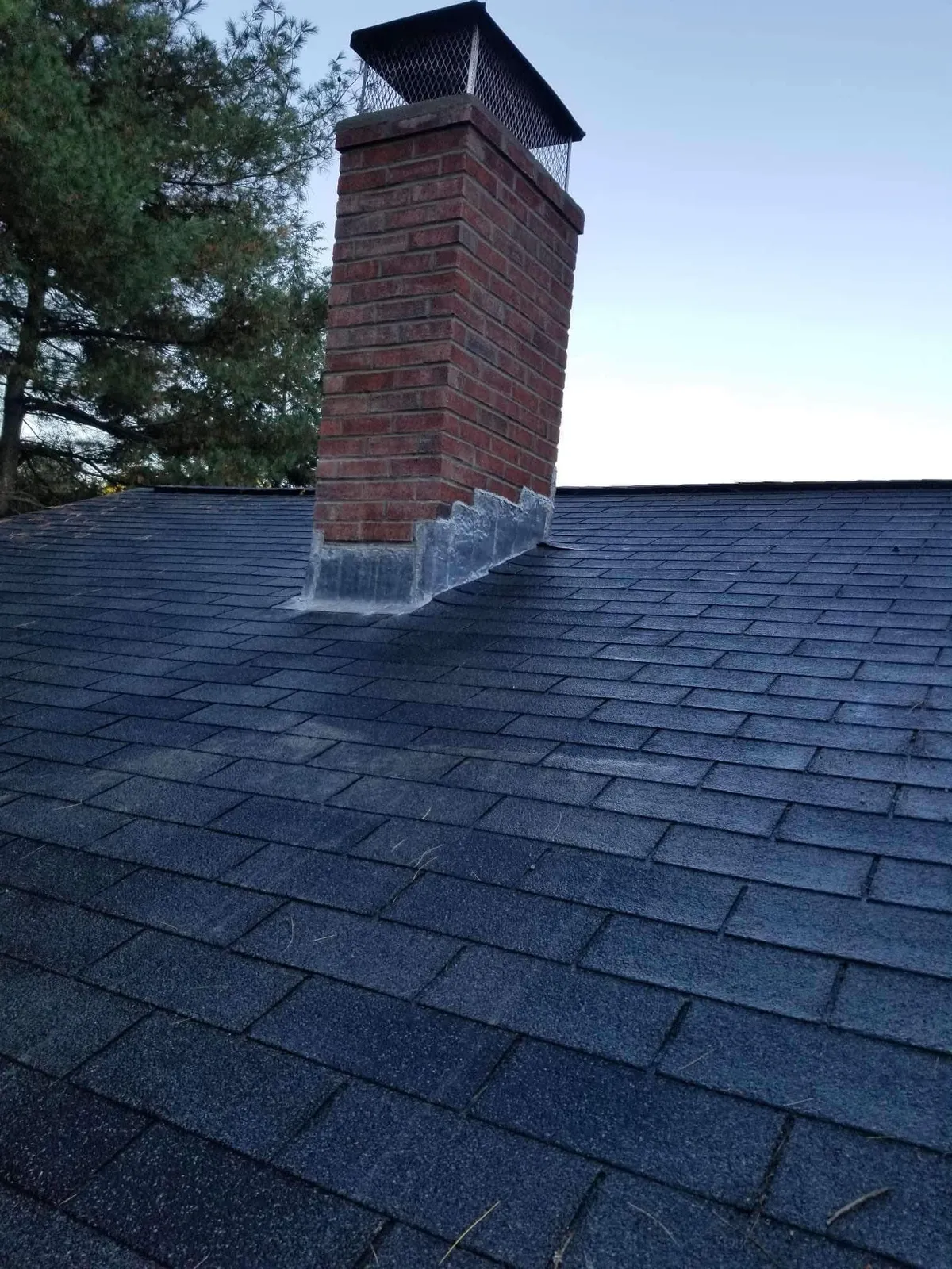 Dark shingled roof with a red brick chimney against a pale evening sky
