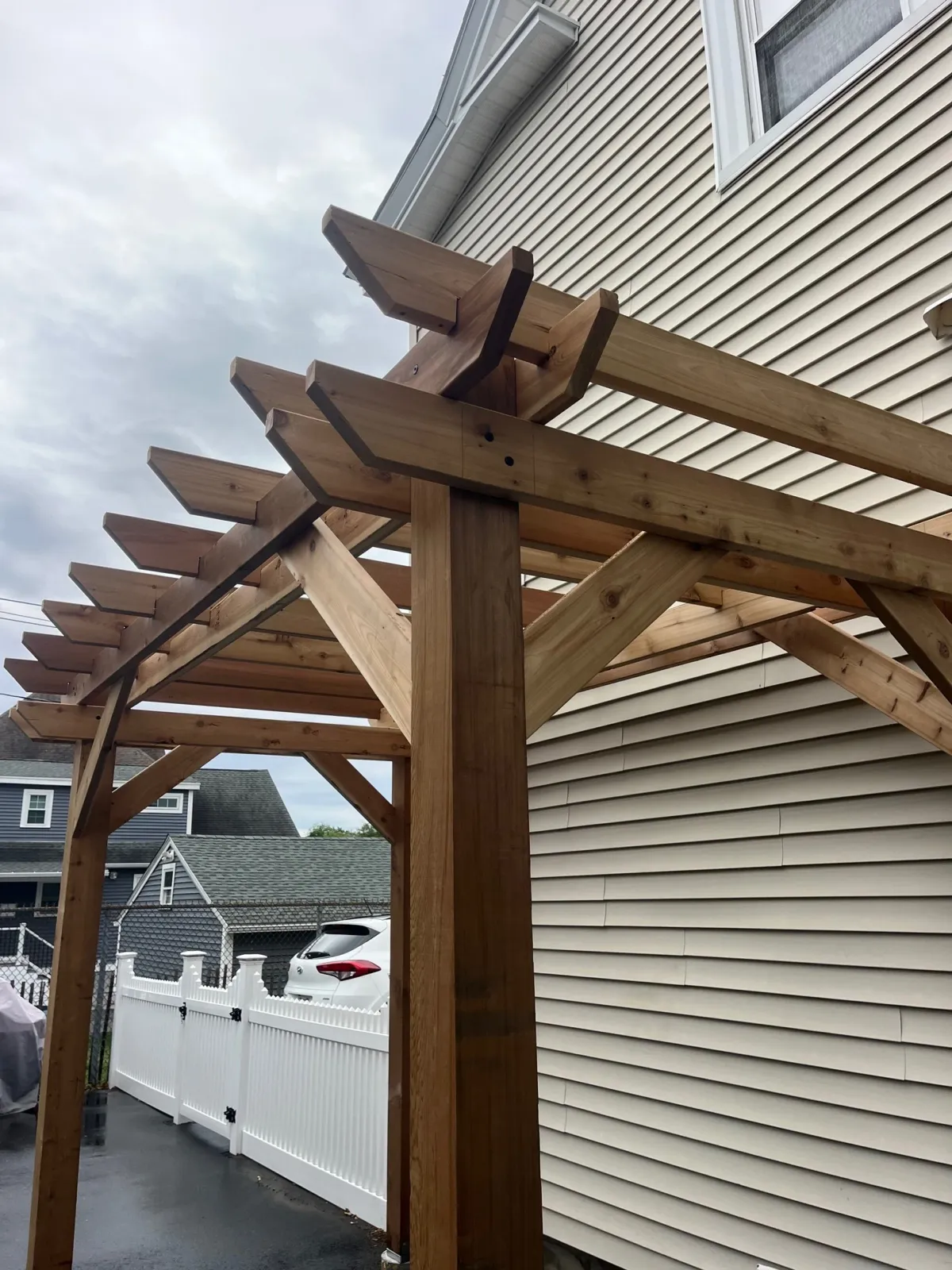 Wooden structure being built next to a house with beige horizontal siding and a window. The structure is supported by vertical posts and has cross beams.