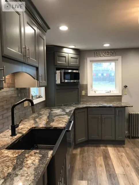 Modern kitchen with gray cabinets, granite countertops, brick backsplash, black sink, microwave, window with 'BAKERY' sign above, and wooden flooring.