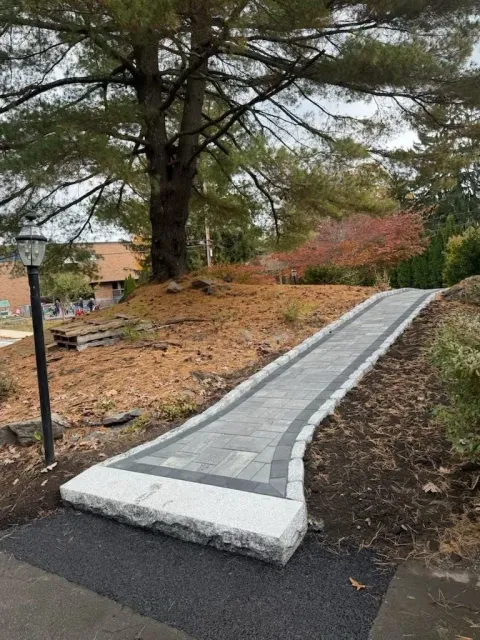 A paved stone walkway with granite curbing leads uphill through a wooded area near a street lamp.