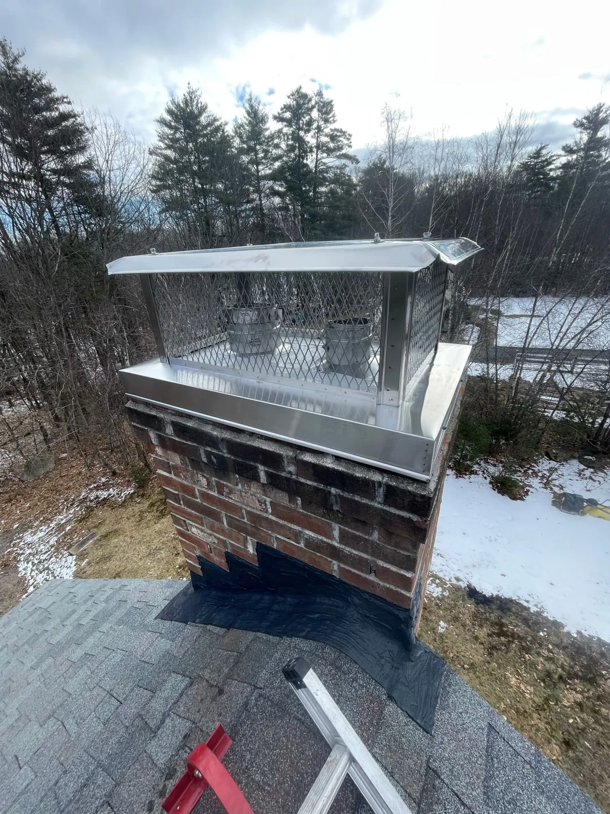 A chimney with a metal cap and vent contains two propane tanks, situated on a rooftop with shingles, overlooking a wooded area with snow on the ground.