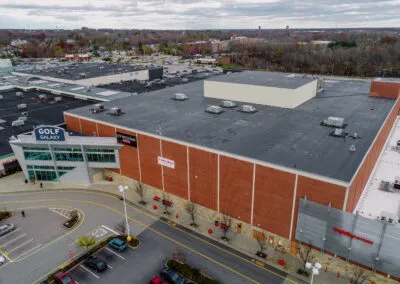 Aerial view of a large shopping mall with a brick exterior and flat black roof. Signs for Target and Golf Galaxy are visible. The parking lot has a few cars and yellow-lined roads, with trees and a cloudy sky in the background.