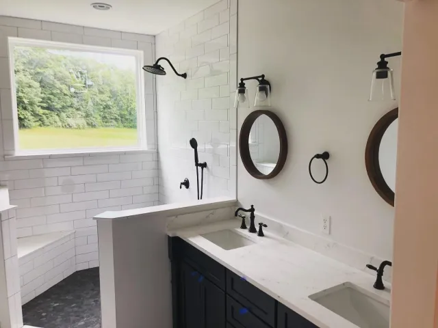 Modern bathroom with white tiled walls, a large window, a double vanity with white marble top, black fixtures, two round mirrors with dark wood frames, and a walk-in shower with black fixtures and a window showing greenery outside.