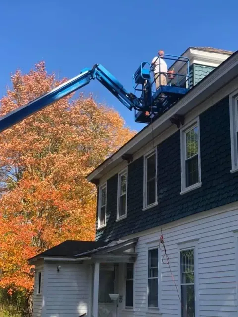 Worker in a blue lift repairs a roof beside a house with white siding and dark trim in autumn.