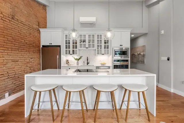 Modern kitchen with white cabinets, an island with a white countertop, four wooden bar stools, a brick accent wall, and stainless steel appliances.