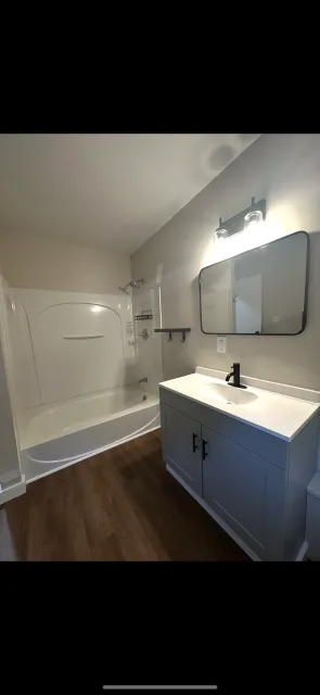Bathroom with a bathtub and shower combination, a small vanity with a mirror, and wooden flooring.