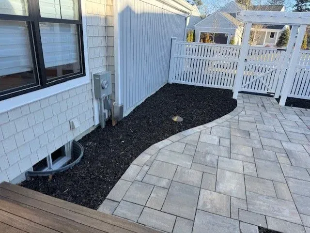 Side yard with a paved patio, a mulched area, and a white picket fence. The house has shingle siding.