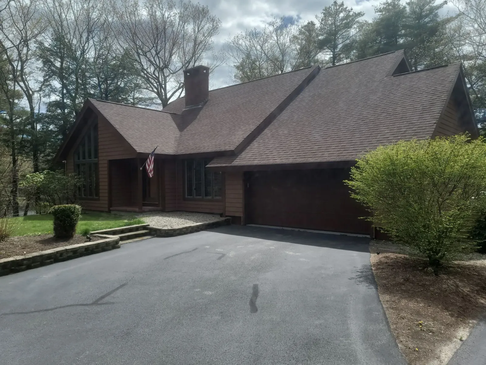 House with brown roof and garage at end of a curved driveway, surrounded by trees and shrubs