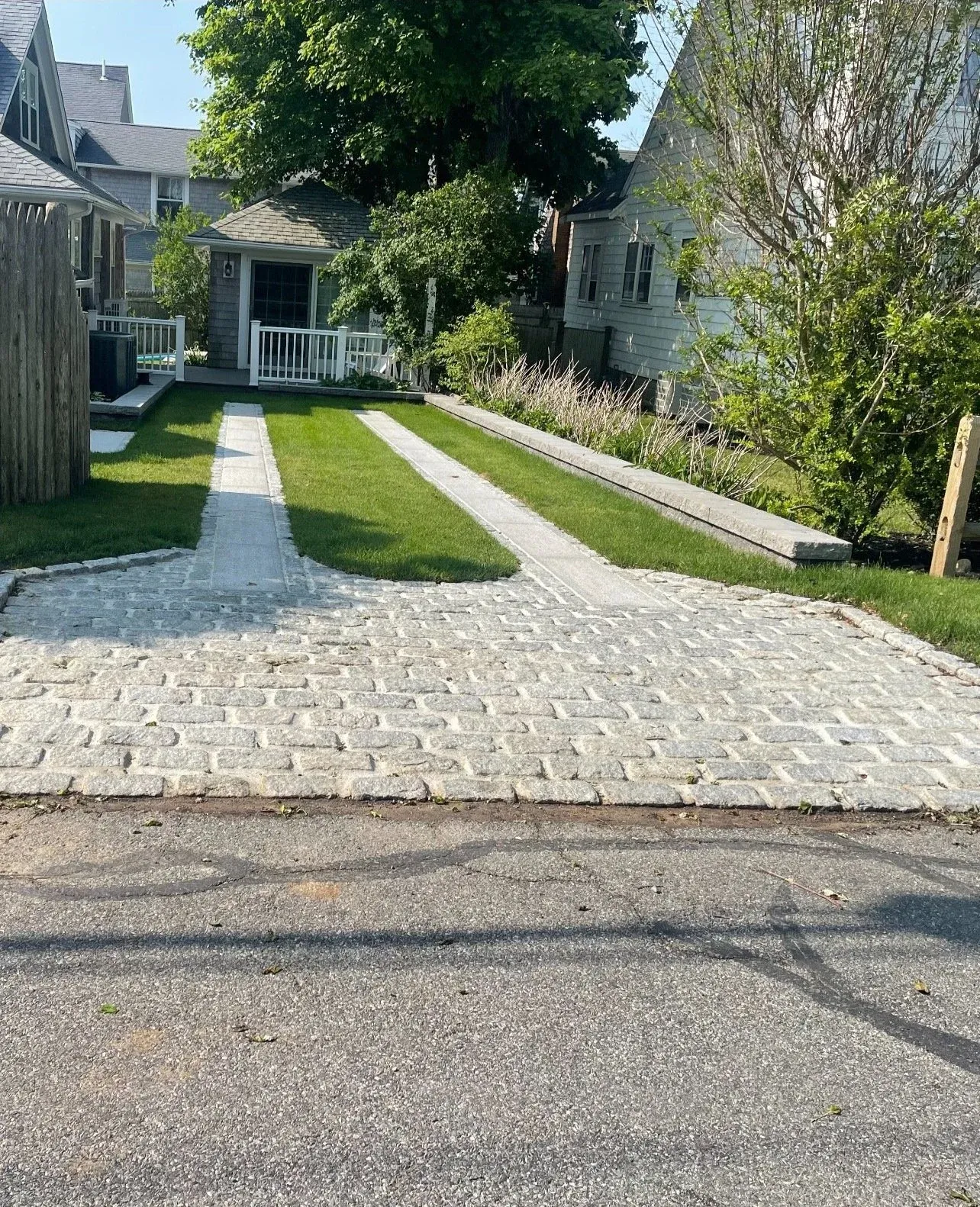 Stone driveway leading to a small house with a white picket fence, surrounded by green grass, trees, and neighboring houses.